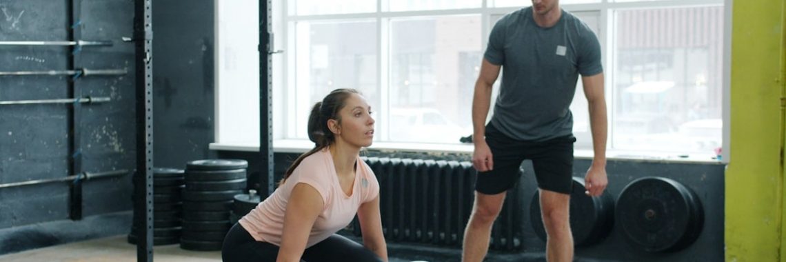 Strong young lady in sportswear is lifting heavy barbell while instructor handsome guy is watching during bodybuilding training in sports gym.